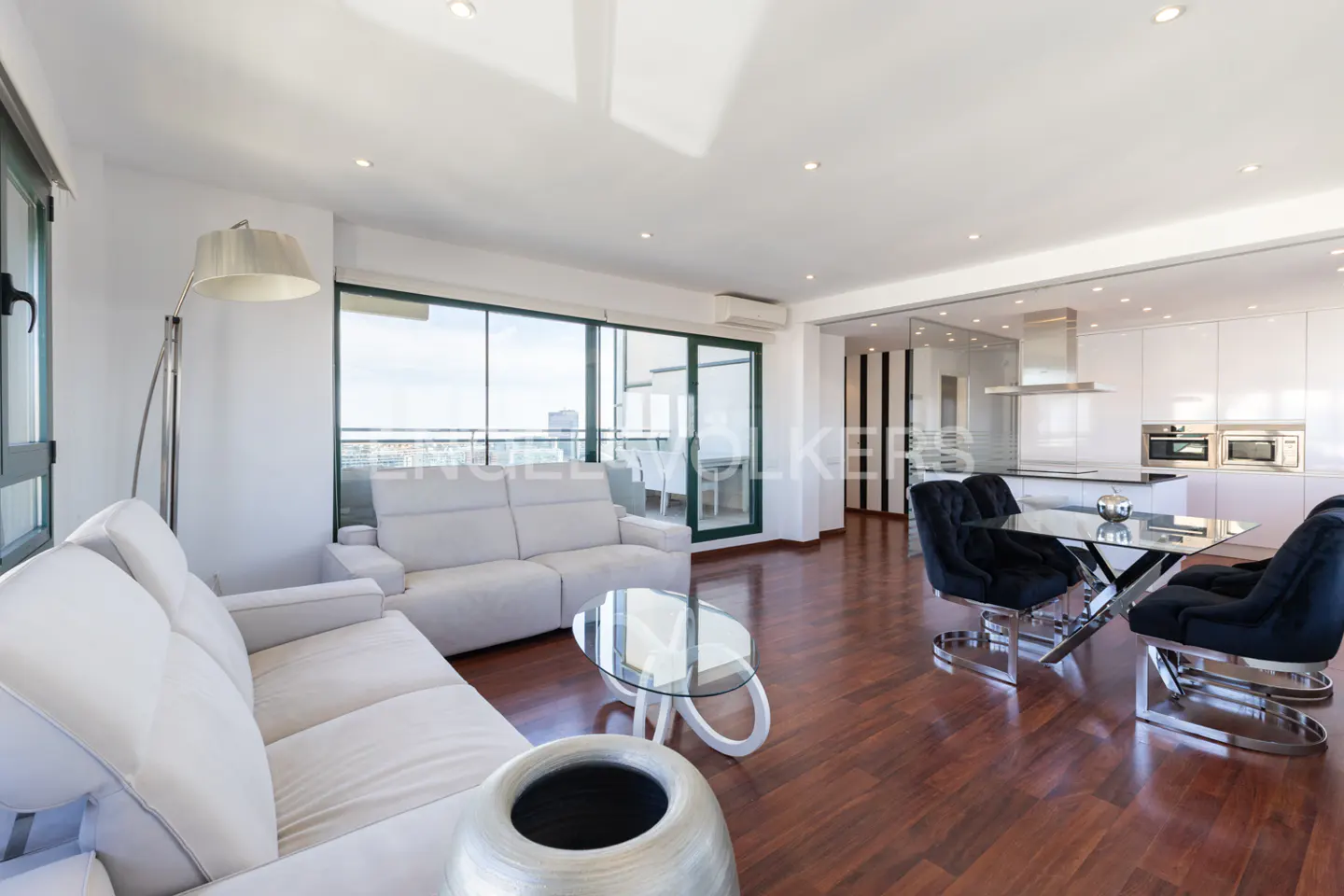 Open-concept living space with white sofas, glass table, and black chairs on a dark wood floor. A modern white kitchen is in the background.