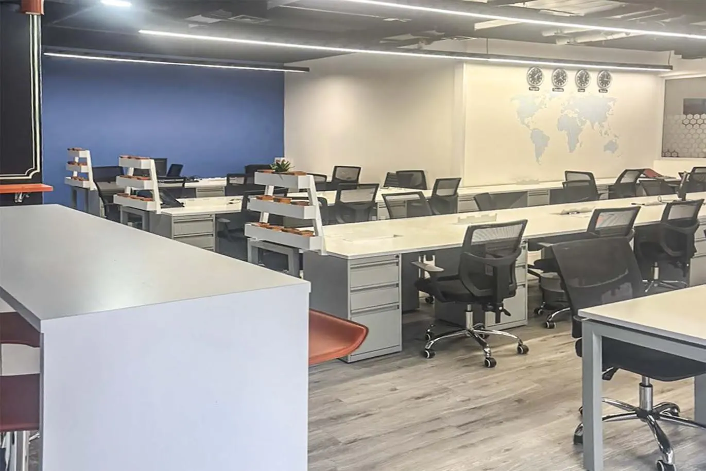 Bright, modern office space with rows of white desks and black mesh chairs. A world map and clocks adorn the wall. Blue accent wall in the background.