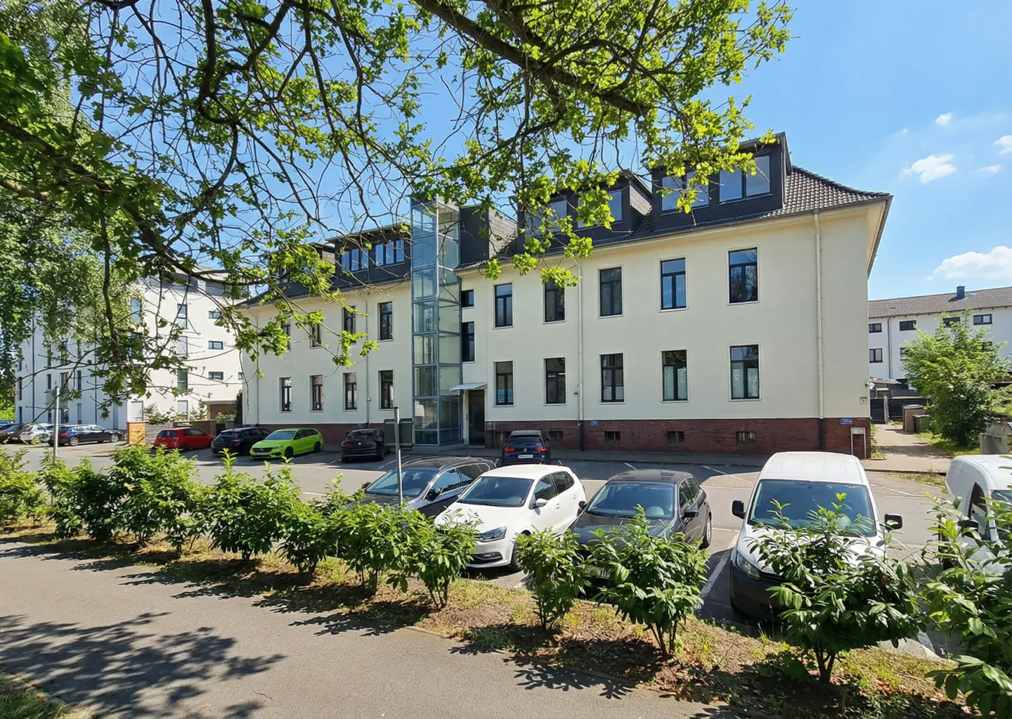 A three-story cream building with a glass elevator and dark trim, cars parked in front.