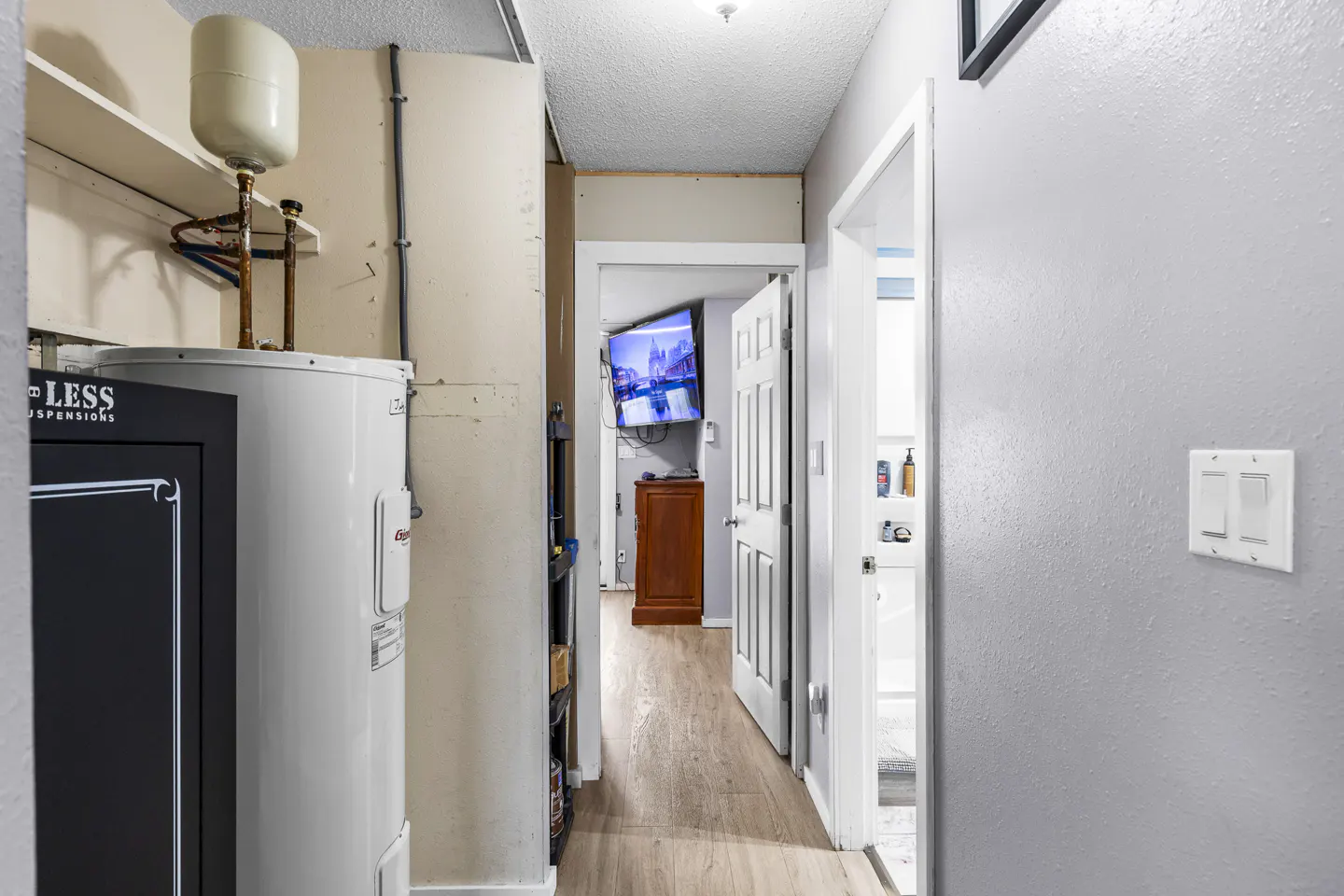Utility area with a water heater and safe, leading to a hallway with a white door and a view into a room with a TV.