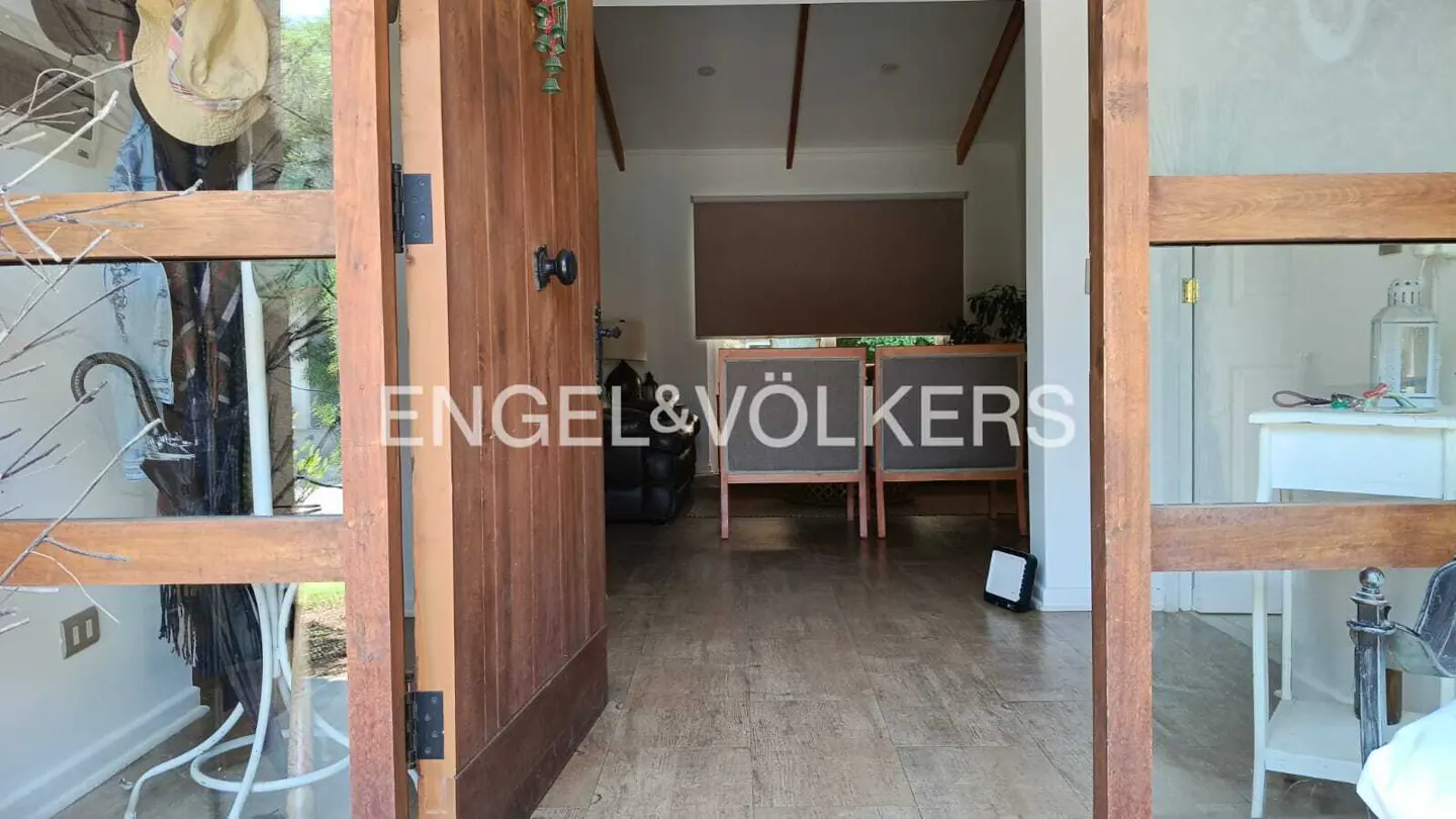 View through an open wooden door into a living room with two grey chairs, wood-look tile floors, and a brown window shade.