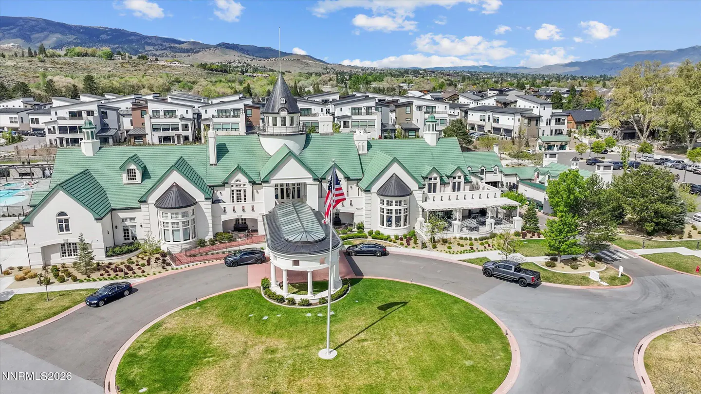 Aerial view of a white clubhouse with a green roof, an American flag, and cars in a circular driveway. Mountains in the background.