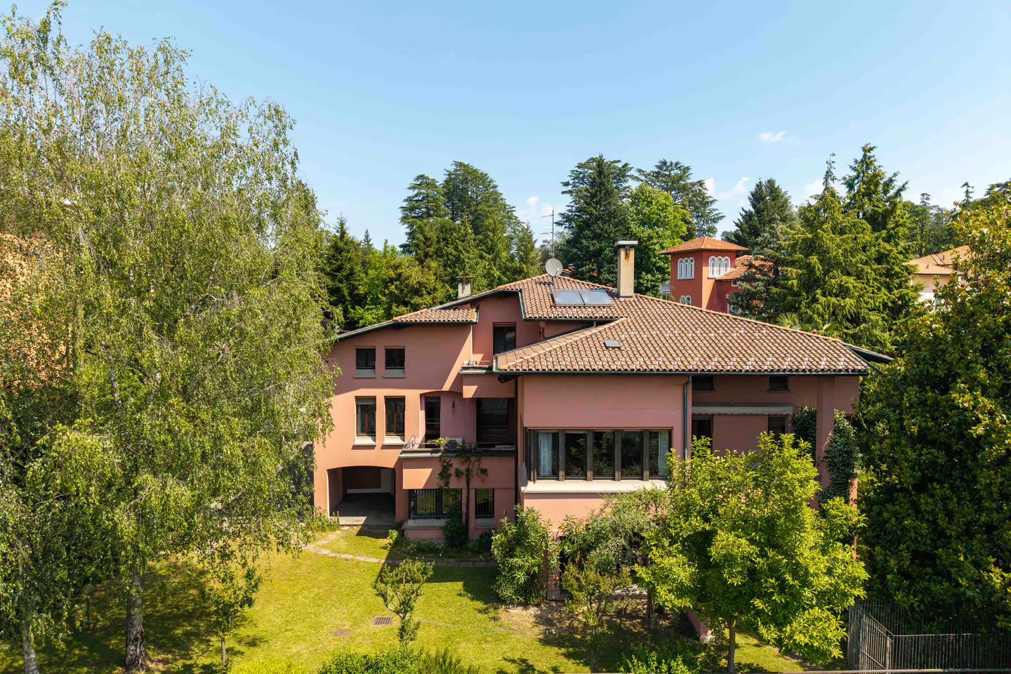 A multi-story pink villa with a tiled roof, surrounded by lush green trees and a lawn under a clear blue sky.
