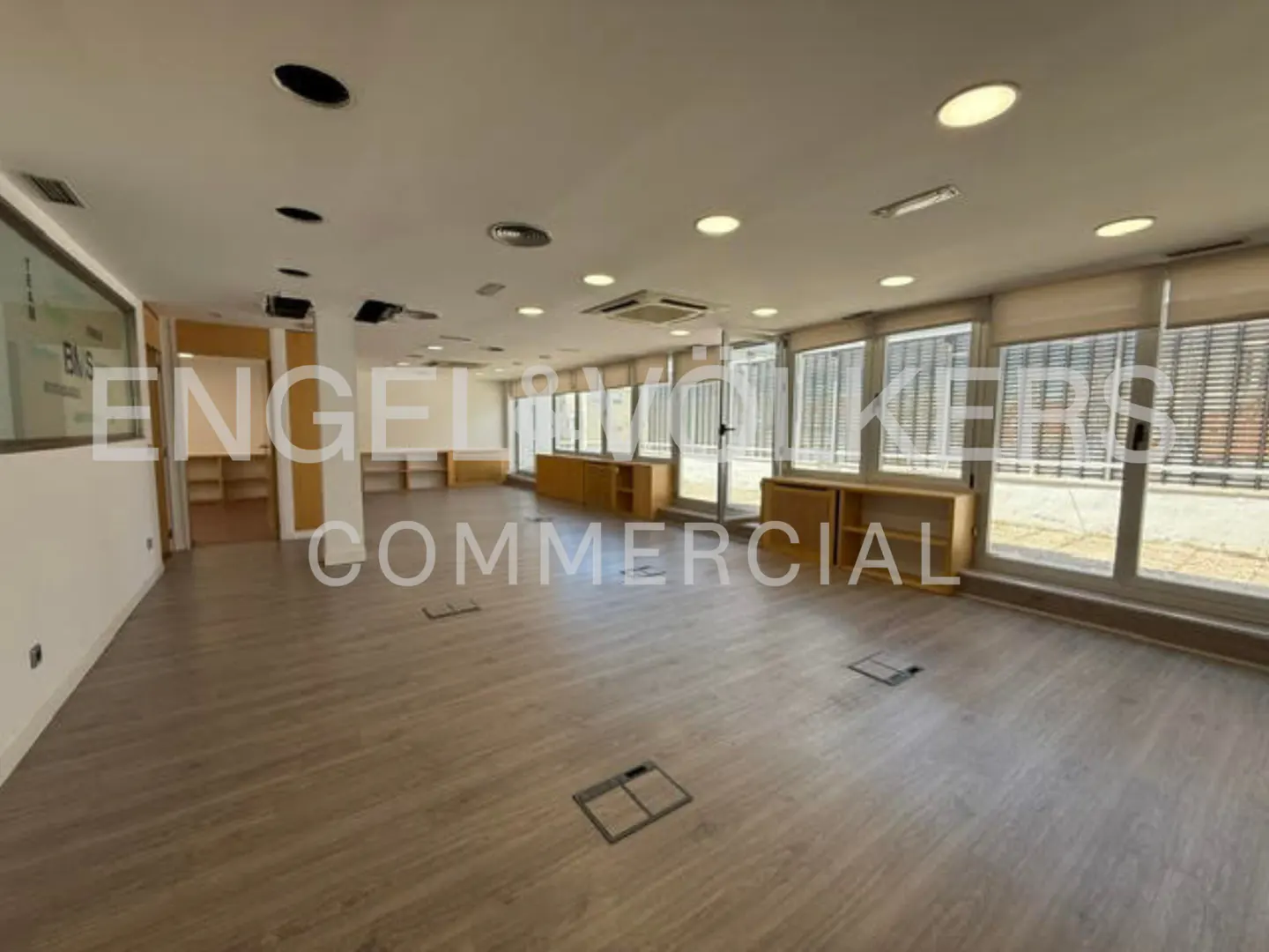Spacious, empty commercial office with light wood floors, recessed ceiling lights, and large windows with blinds.