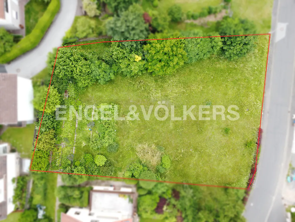 Aerial view of a grassy lot outlined in red, surrounded by trees and houses. "Engel & Völkers" logo is superimposed.