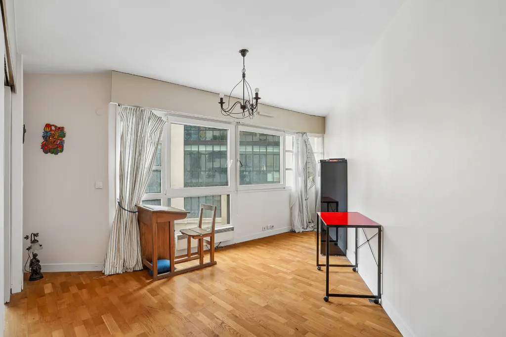 Bright room with wood floors, large window, desk, chair, and red table. White walls and curtains. Chandelier hangs from the ceiling.