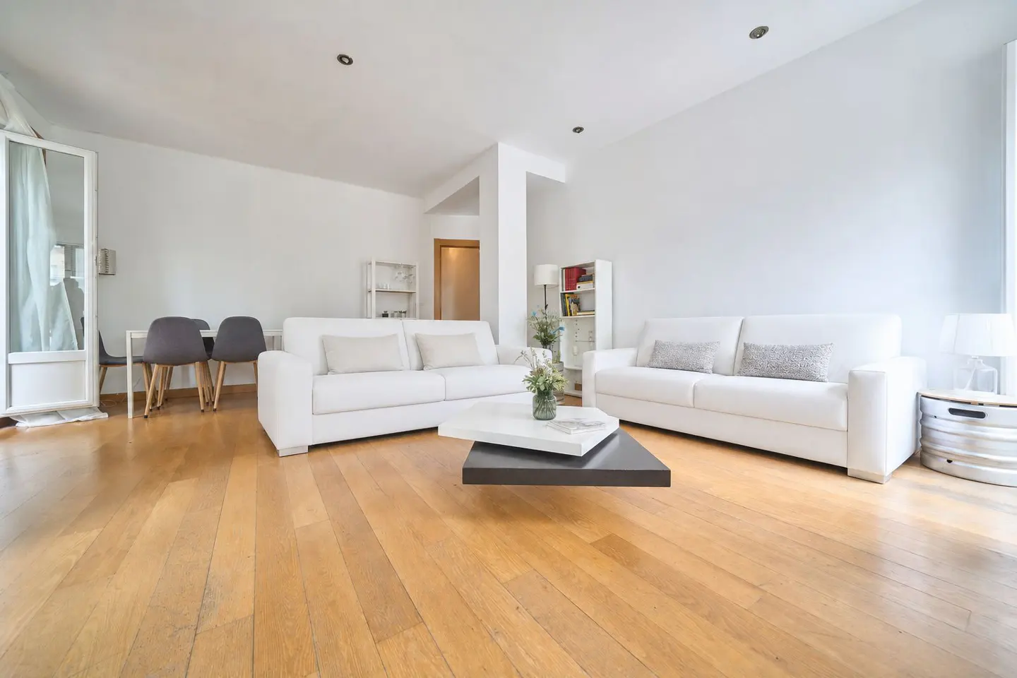 Bright living room with hardwood floors, white walls, and two white sofas. A modern coffee table sits in the center. Dining area visible in the background.