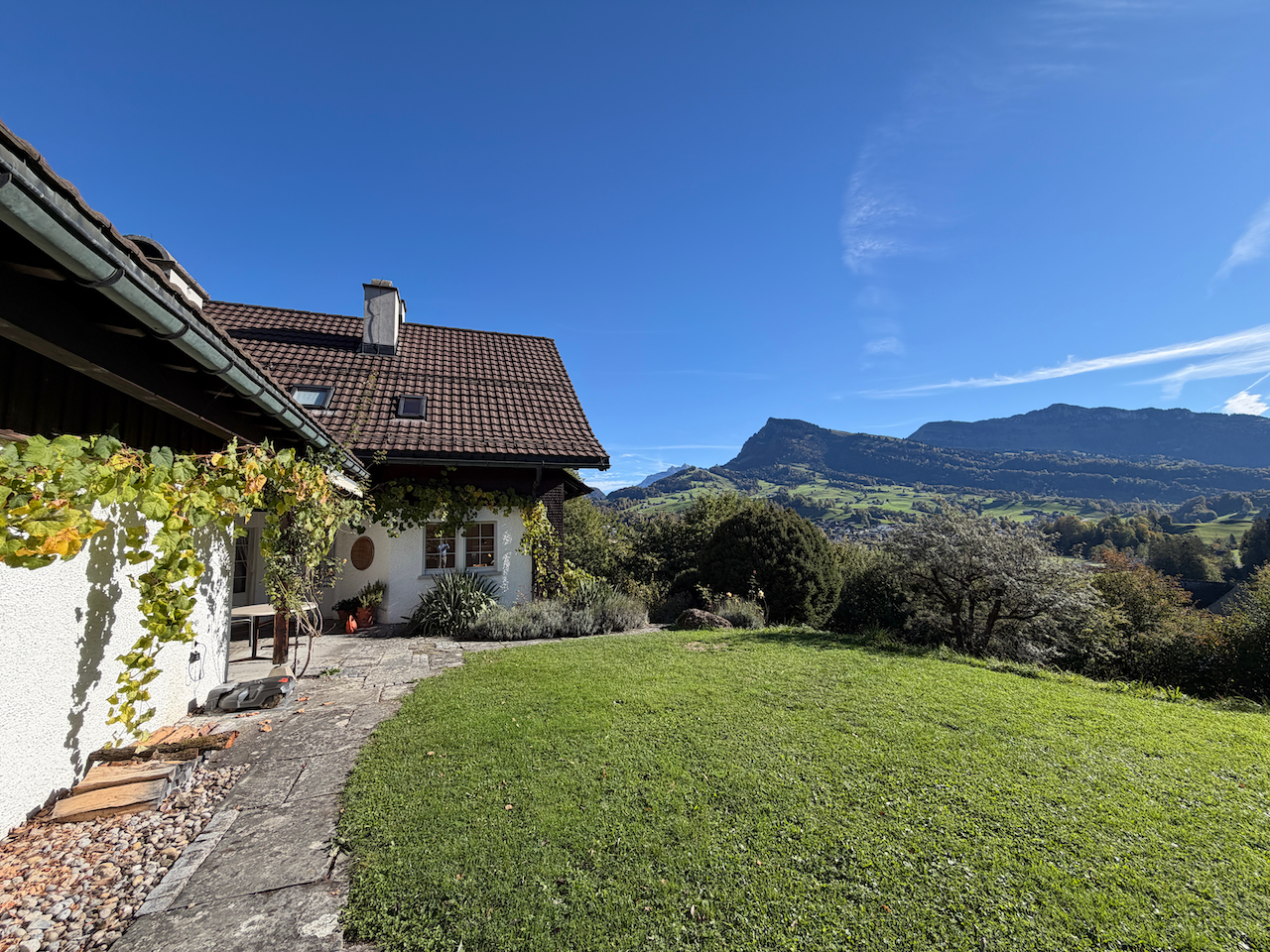 A house with a brown tiled roof and white walls overlooks a green lawn and a scenic mountain landscape under a clear blue sky. [AI-generated text]