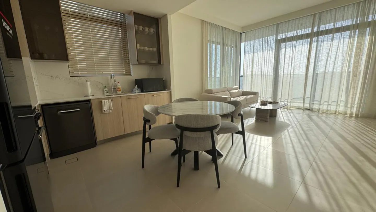 Modern open-plan kitchen and living area with a round dining table, light wood cabinets, and floor-to-ceiling curtains.