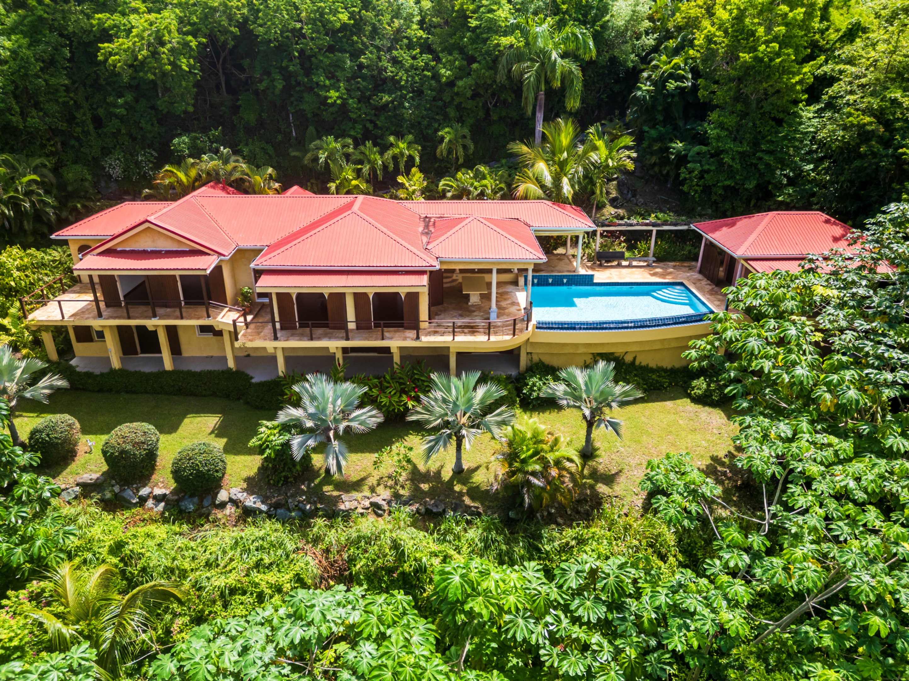 Aerial view of a yellow two-story house with a red roof and a blue pool surrounded by lush green trees.