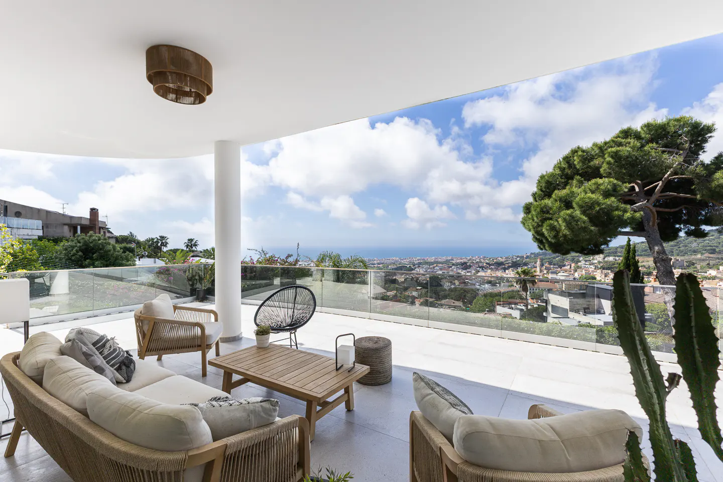 Bright, covered patio with wicker furniture, a wood table, and a black chair. A glass railing overlooks a city, ocean, and blue sky.