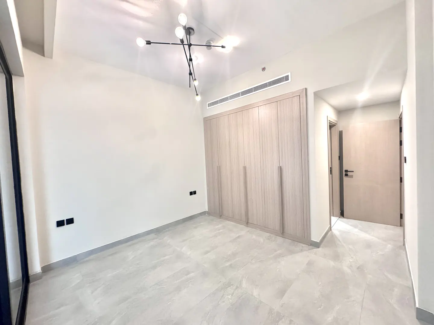 Bright, empty bedroom with light gray tile floor, white walls, and a large, light wood closet. A modern light fixture hangs from the ceiling.