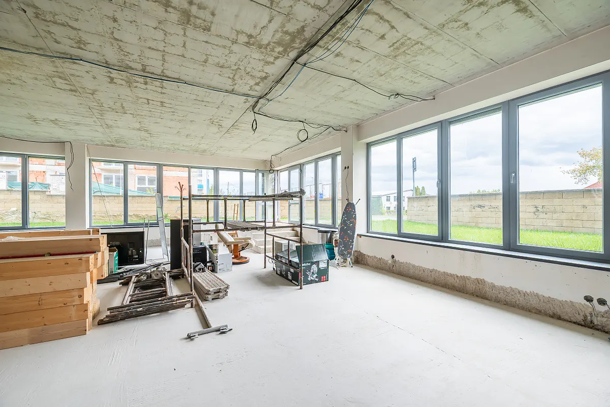 Unfinished room with concrete ceiling, white walls, and large gray windows. Construction materials and equipment are scattered throughout.