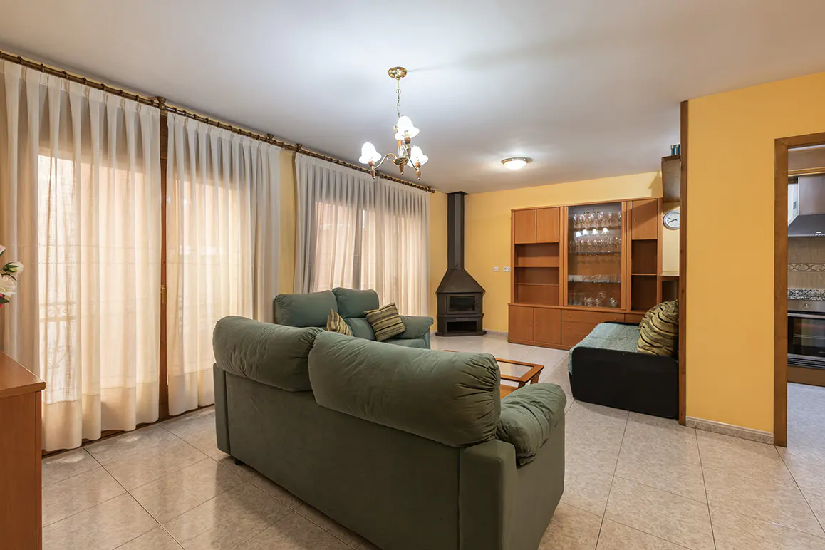 Living room with yellow walls, tile floors, and a green sofa. A fireplace and wood cabinet are against the wall. Curtains cover the windows.
