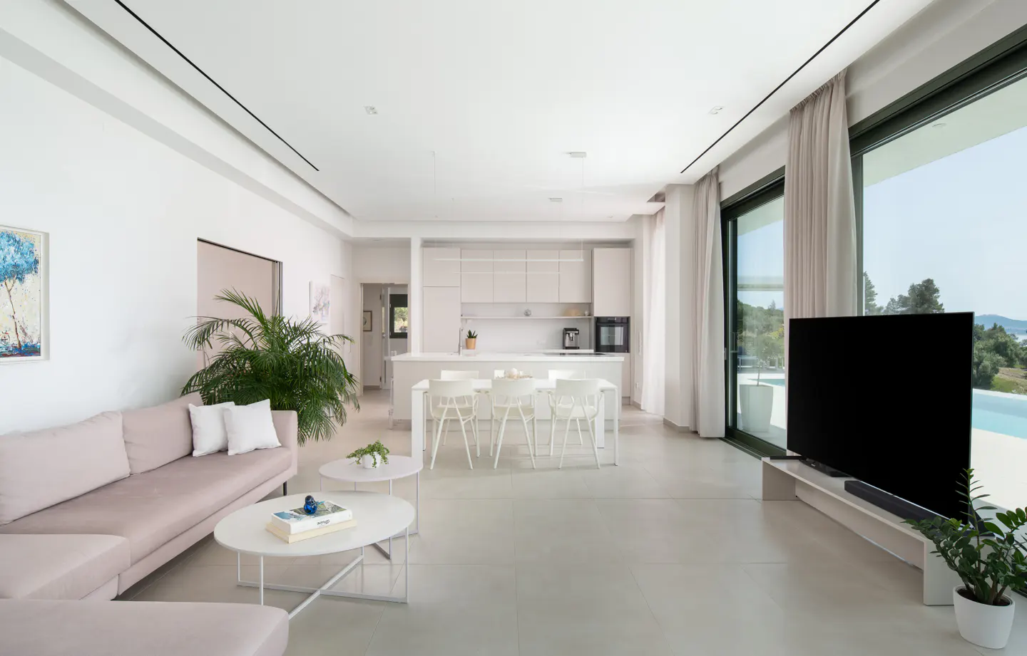 Bright, open-concept living space with a pink sofa, white kitchen, dining table, and large windows overlooking a pool and trees.