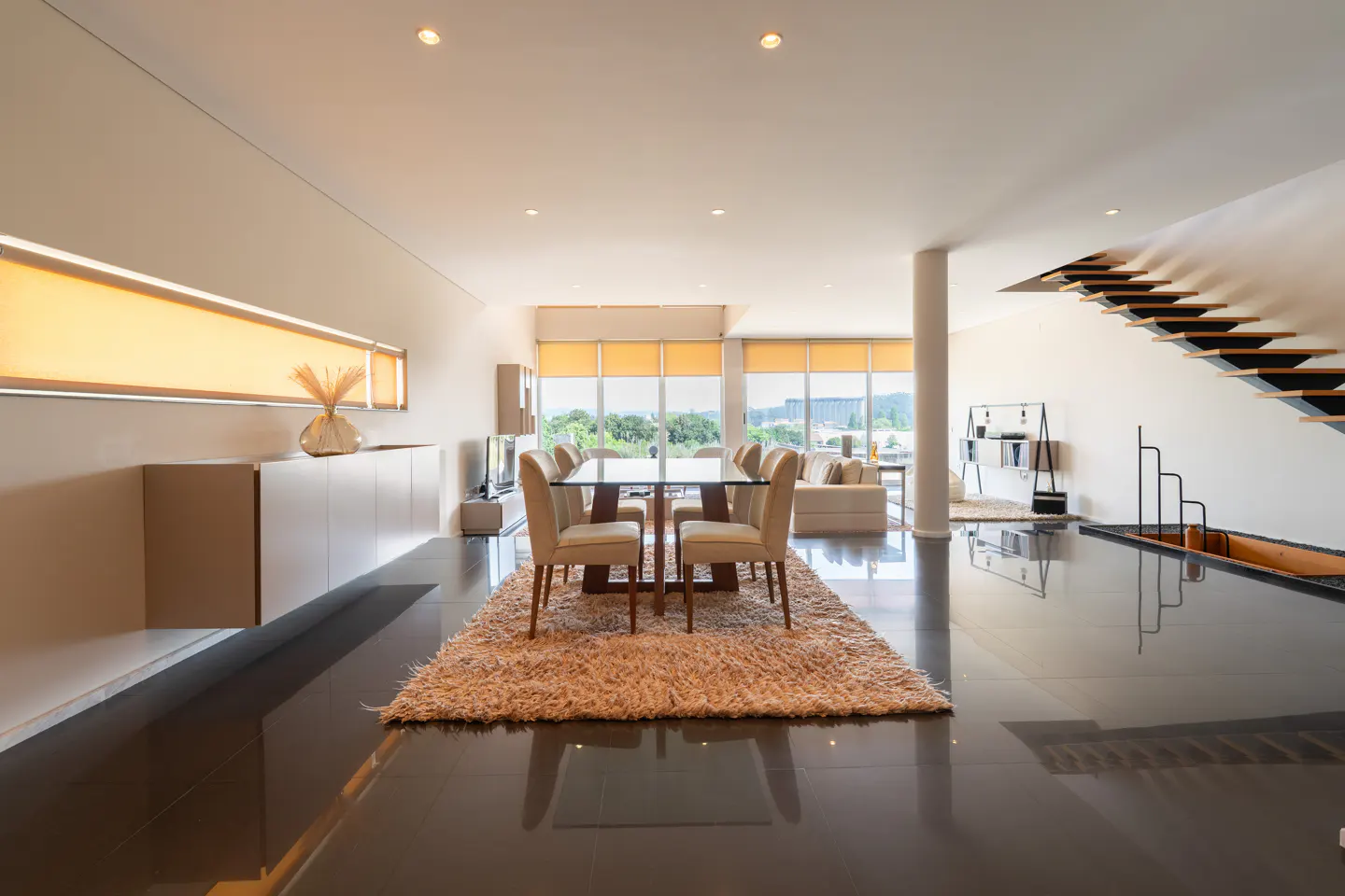 Open-concept living space with a glass dining table, beige chairs, and a shaggy rug on a dark tile floor. Stairs and a living room are visible.