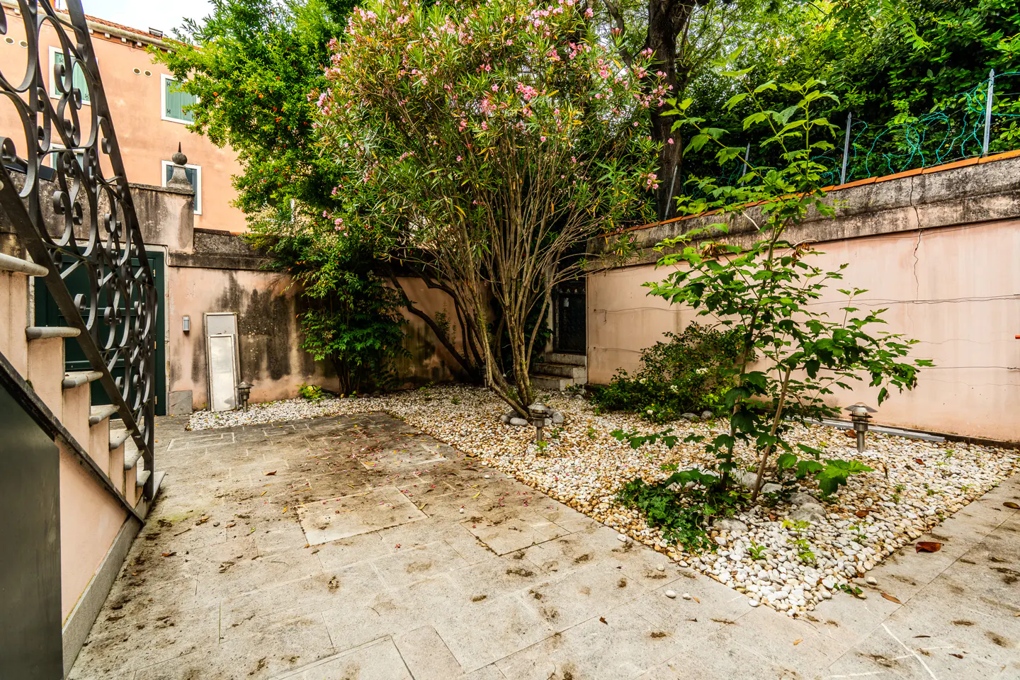 A stone patio with a gravel garden, a flowering tree, and a pink wall, viewed from a black wrought-iron staircase.