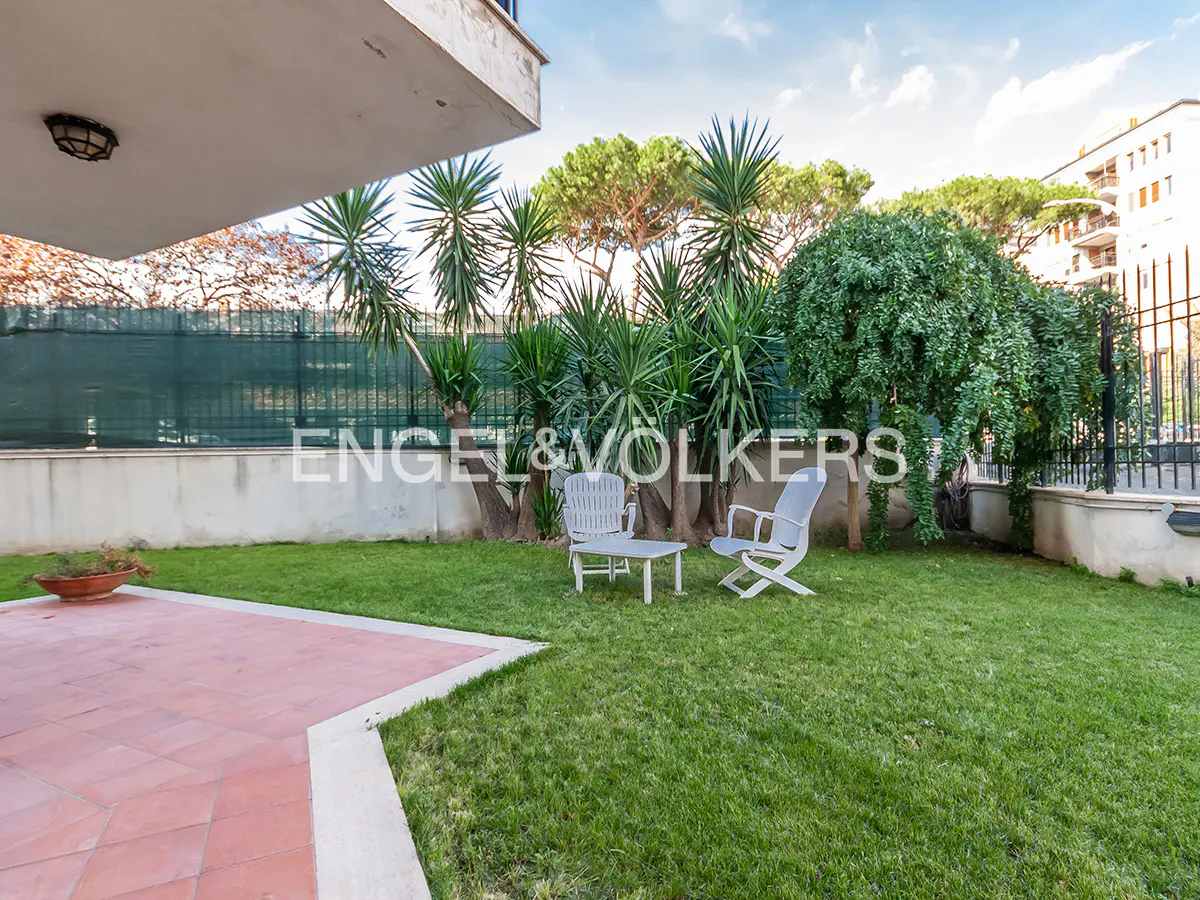A backyard with green grass, palm trees, and a white table and chairs set. A red tiled patio is in the foreground.