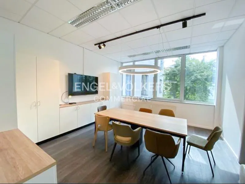 Bright office space with a long wooden table surrounded by chairs, white cabinets, and a large window.