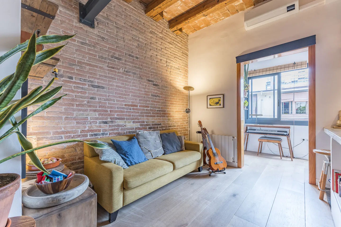 Living room with exposed brick wall, mustard sofa, wooden floors, and a sunlit alcove featuring a keyboard and stool.