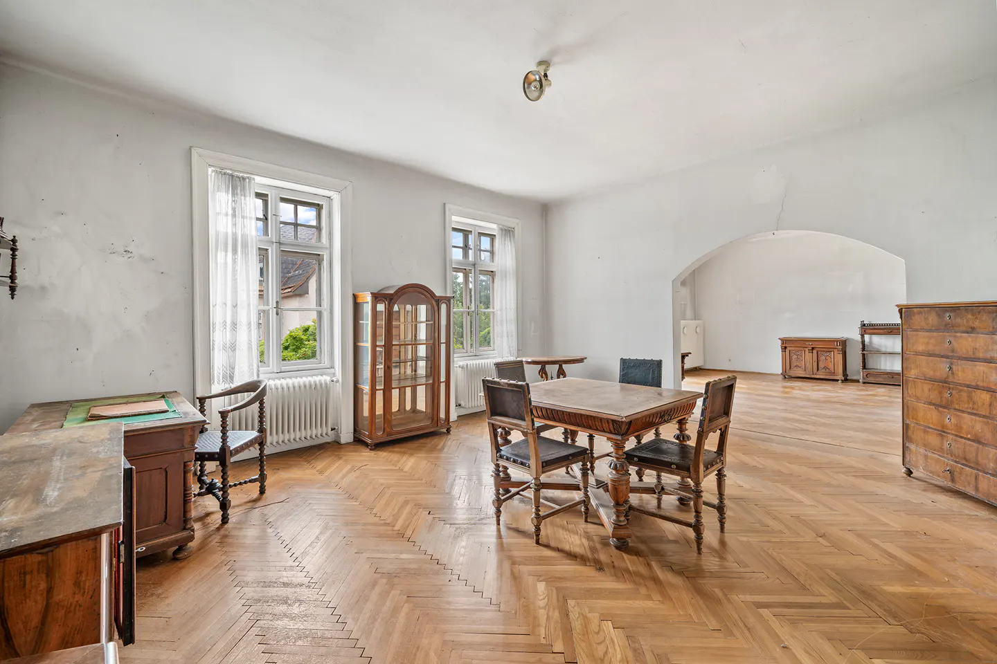 A bright room with white walls, a herringbone floor, and antique wooden furniture. Windows with white frames let in natural light.