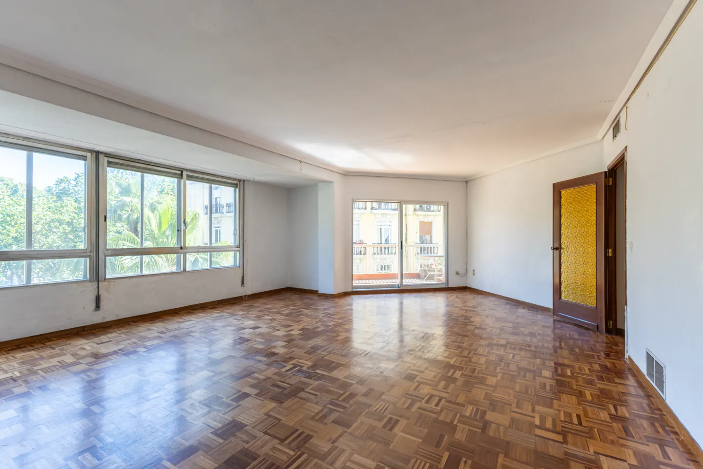 Bright, empty room with parquet floor, white walls, and large windows. A door with textured glass is open. Balcony doors lead to a patio.