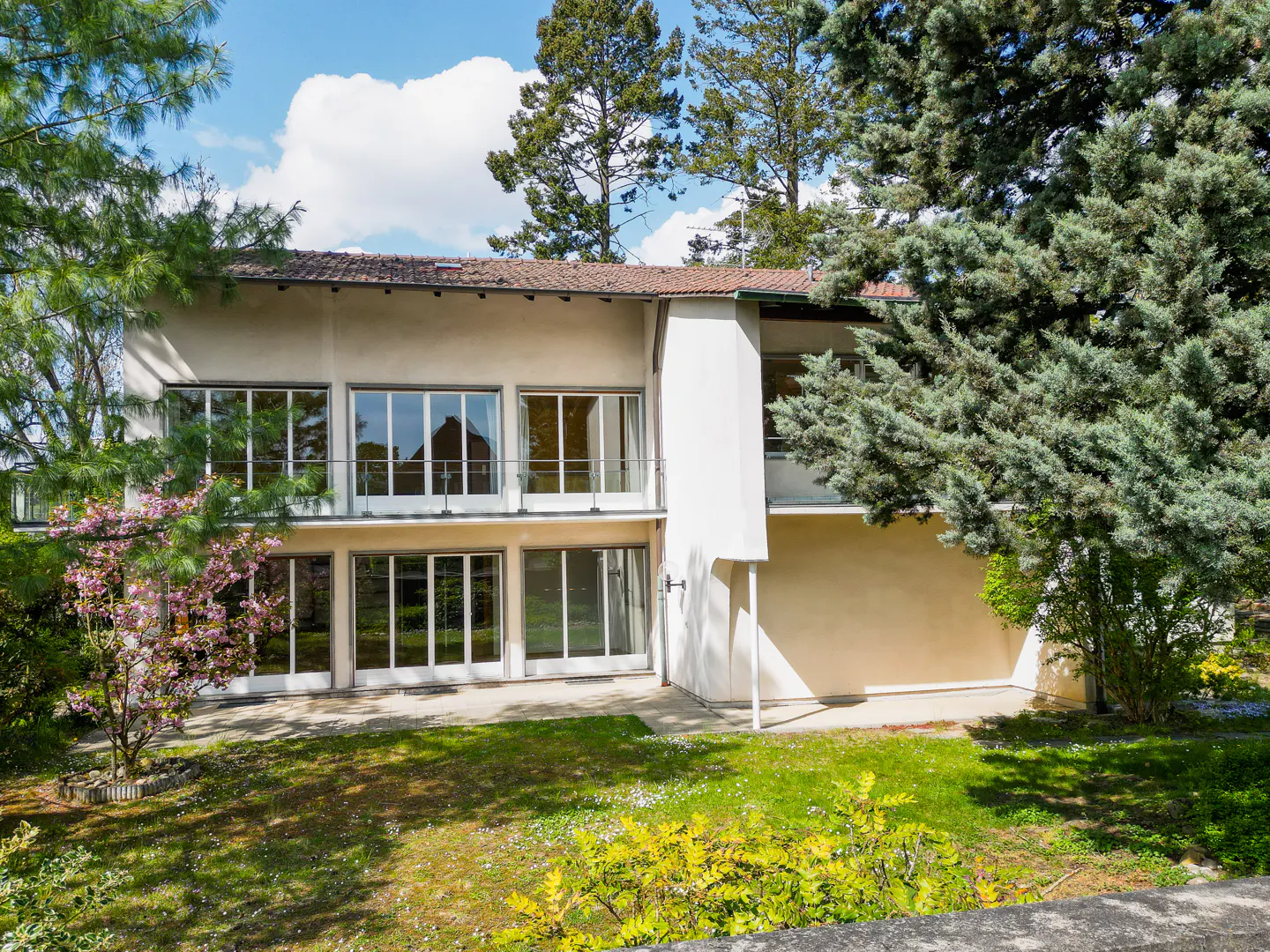 Two-story beige house with large windows and a balcony, surrounded by green trees and a lawn.