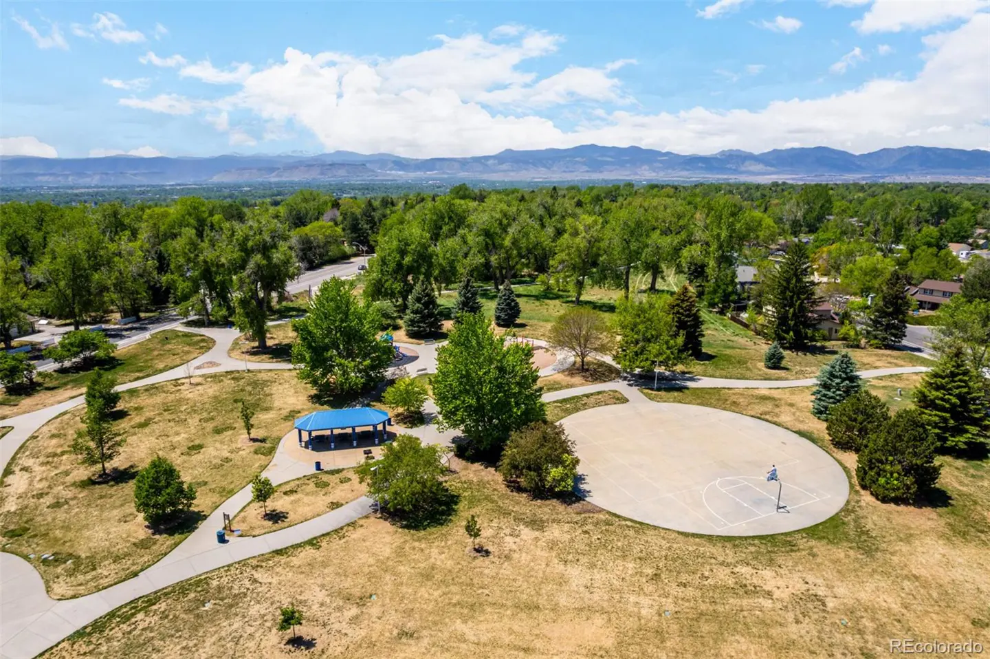 Aerial view of a park with a basketball court, blue picnic shelter, walking paths, trees, and mountains in the background.