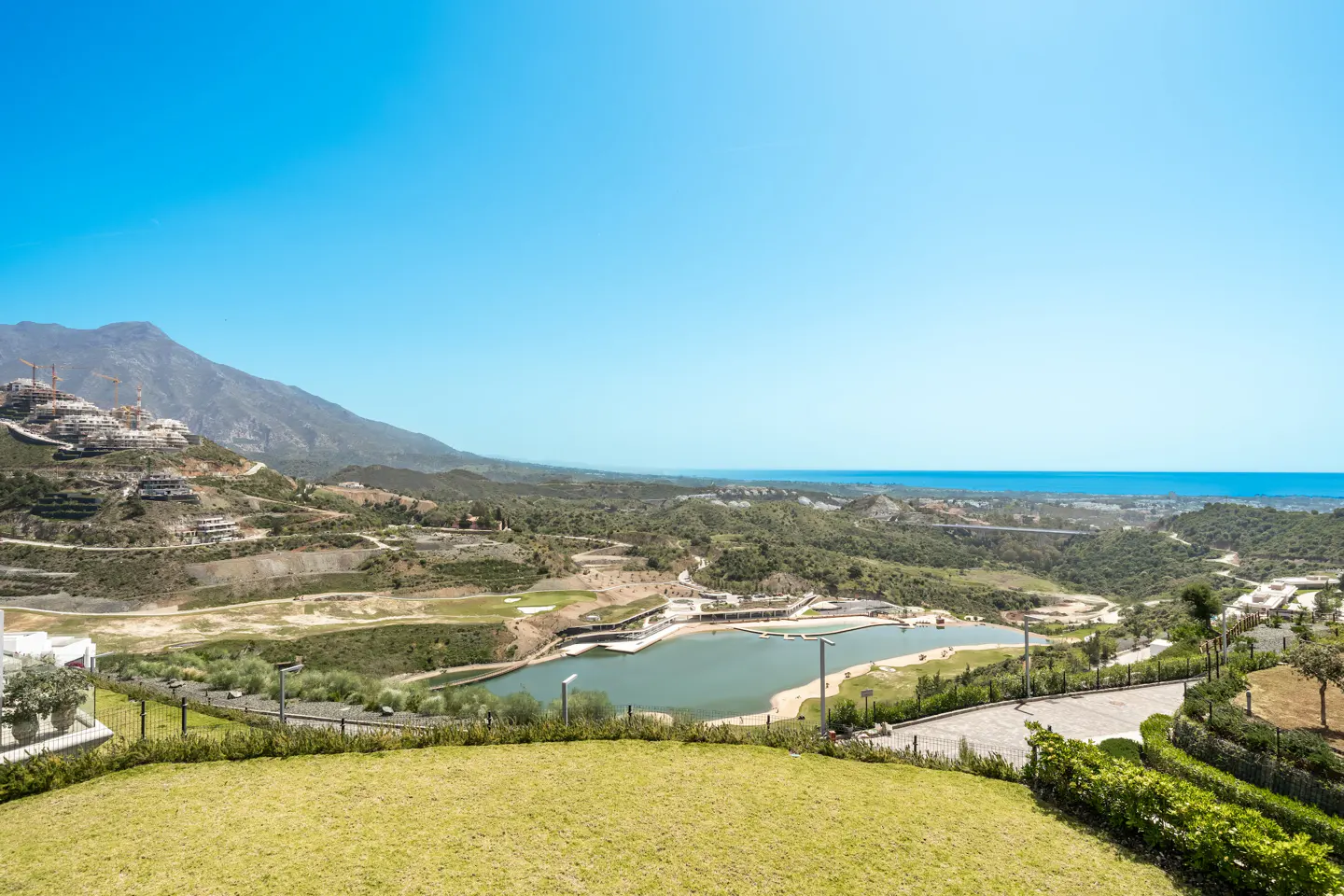 Scenic view of a lake surrounded by green hills, with mountains and construction in the background under a clear blue sky.