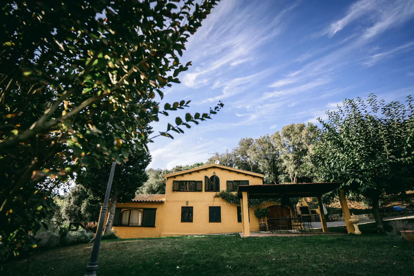 Two-story yellow house with a brown tile roof, dark shutters, and a covered patio, set on a green lawn under a blue sky.