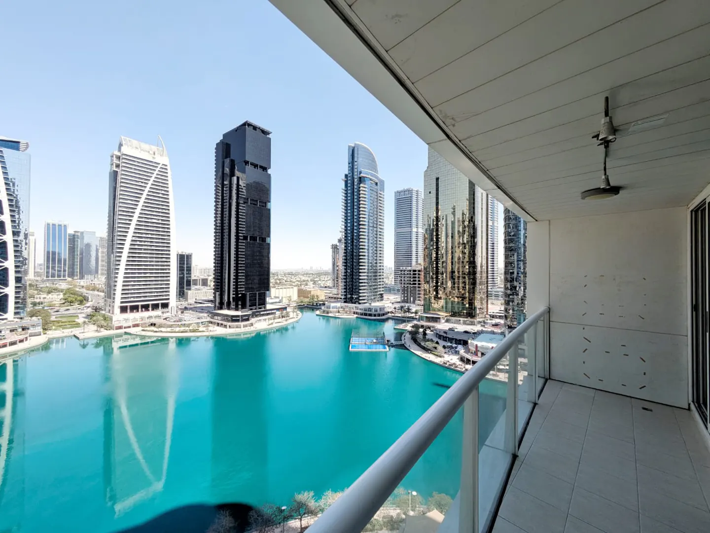 View from a balcony overlooking a bright turquoise lake surrounded by modern skyscrapers under a clear blue sky.