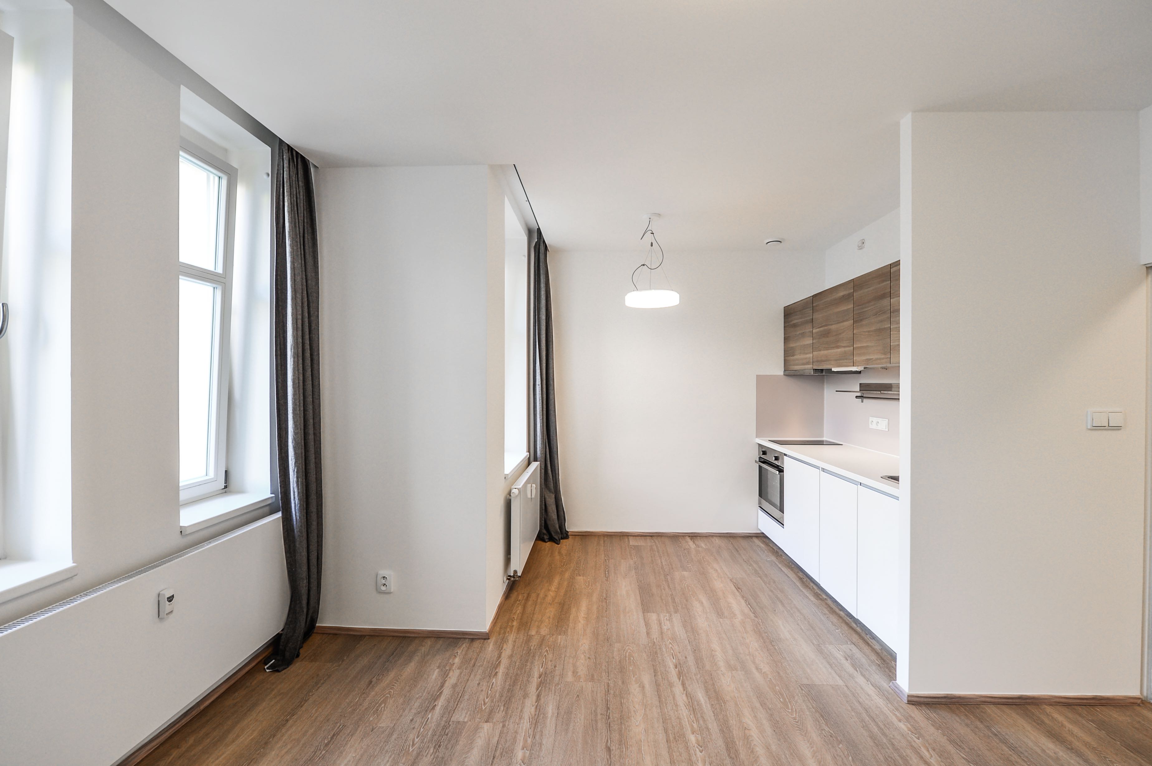 Bright, empty apartment with wood floors, white walls, and a modern kitchen with white cabinets and wood accents. Gray curtains frame the windows.
