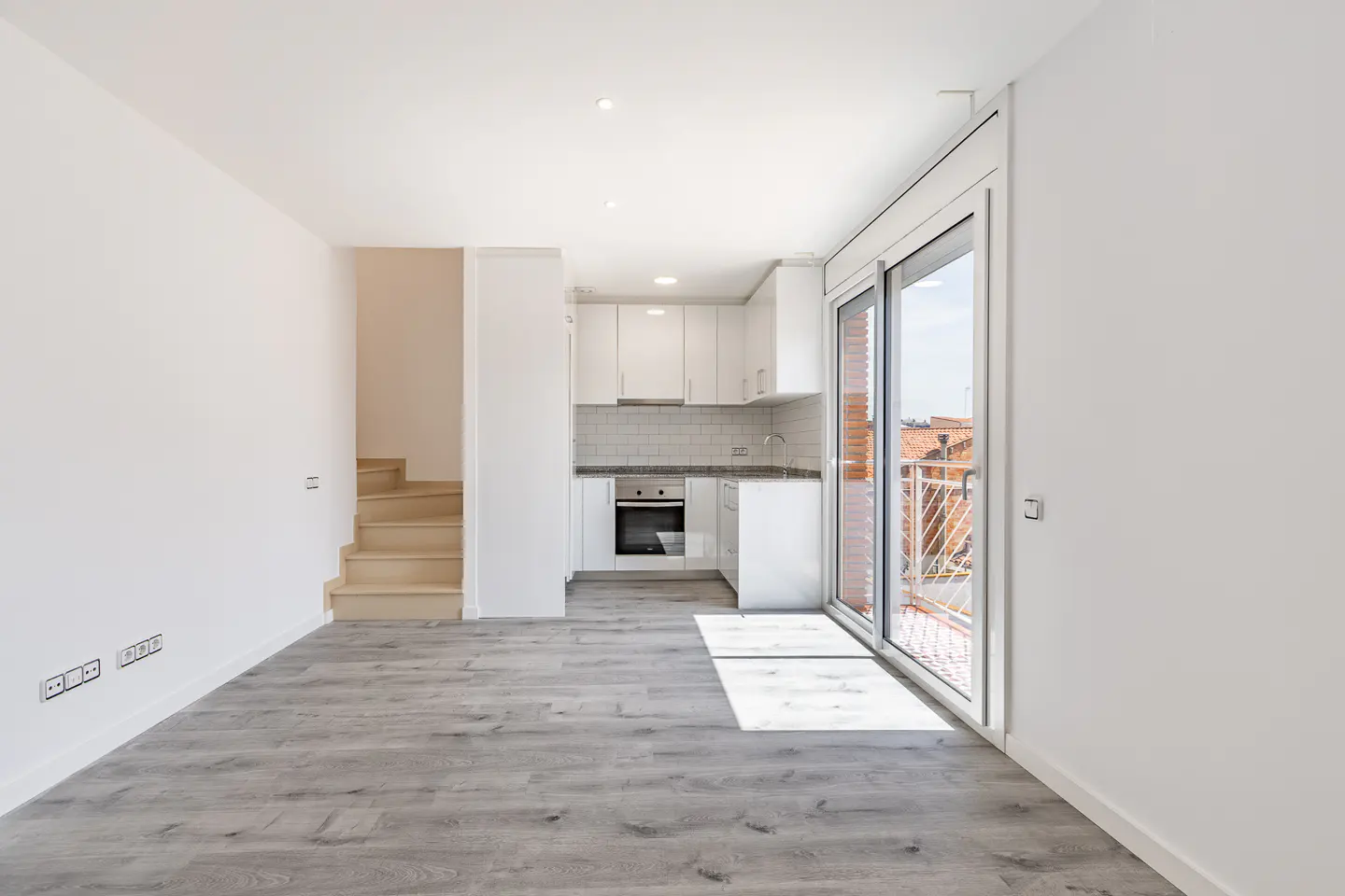Bright, modern apartment interior with gray wood floors, white walls, stairs, and a compact kitchen with white cabinets. Sliding glass doors lead to a balcony.