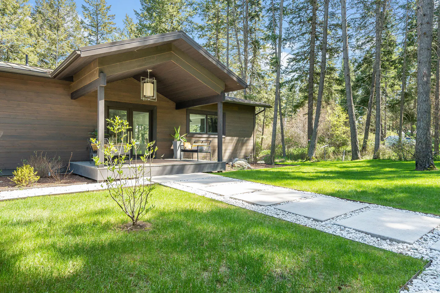 Exterior of a brown, modern home with a stone walkway, green lawn, and tall trees. A covered porch has a hanging lantern and seating.