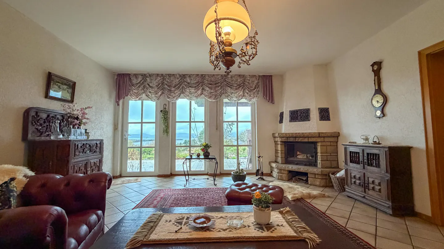 Cozy living room with a stone fireplace, antique furniture, and a view through large windows. A vintage chandelier hangs from the ceiling.