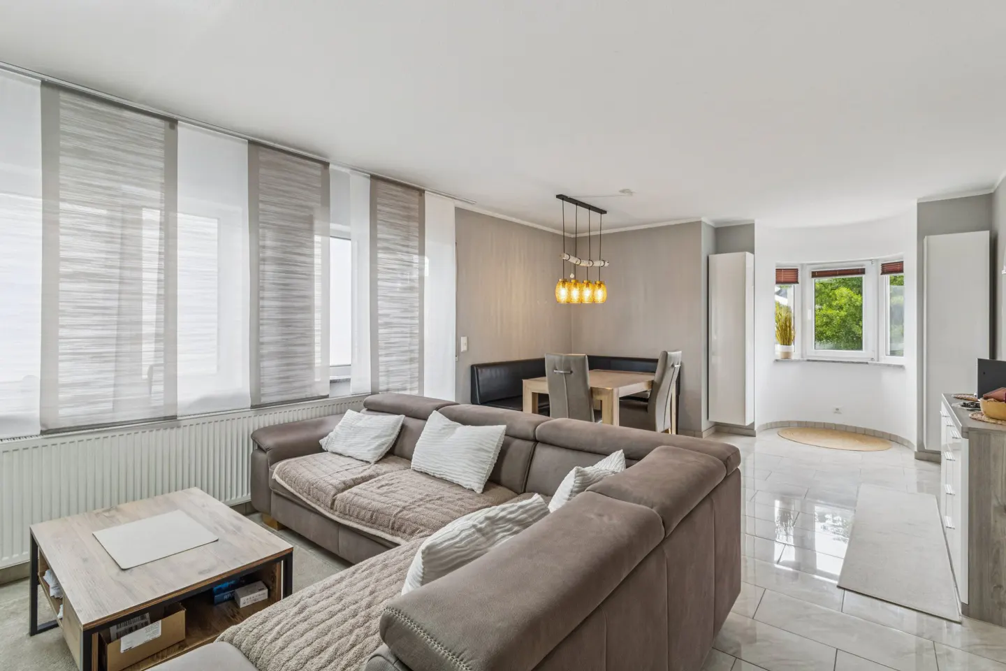 Modern living room with a large brown sectional sofa, wooden coffee table, and a dining area with pendant lights.