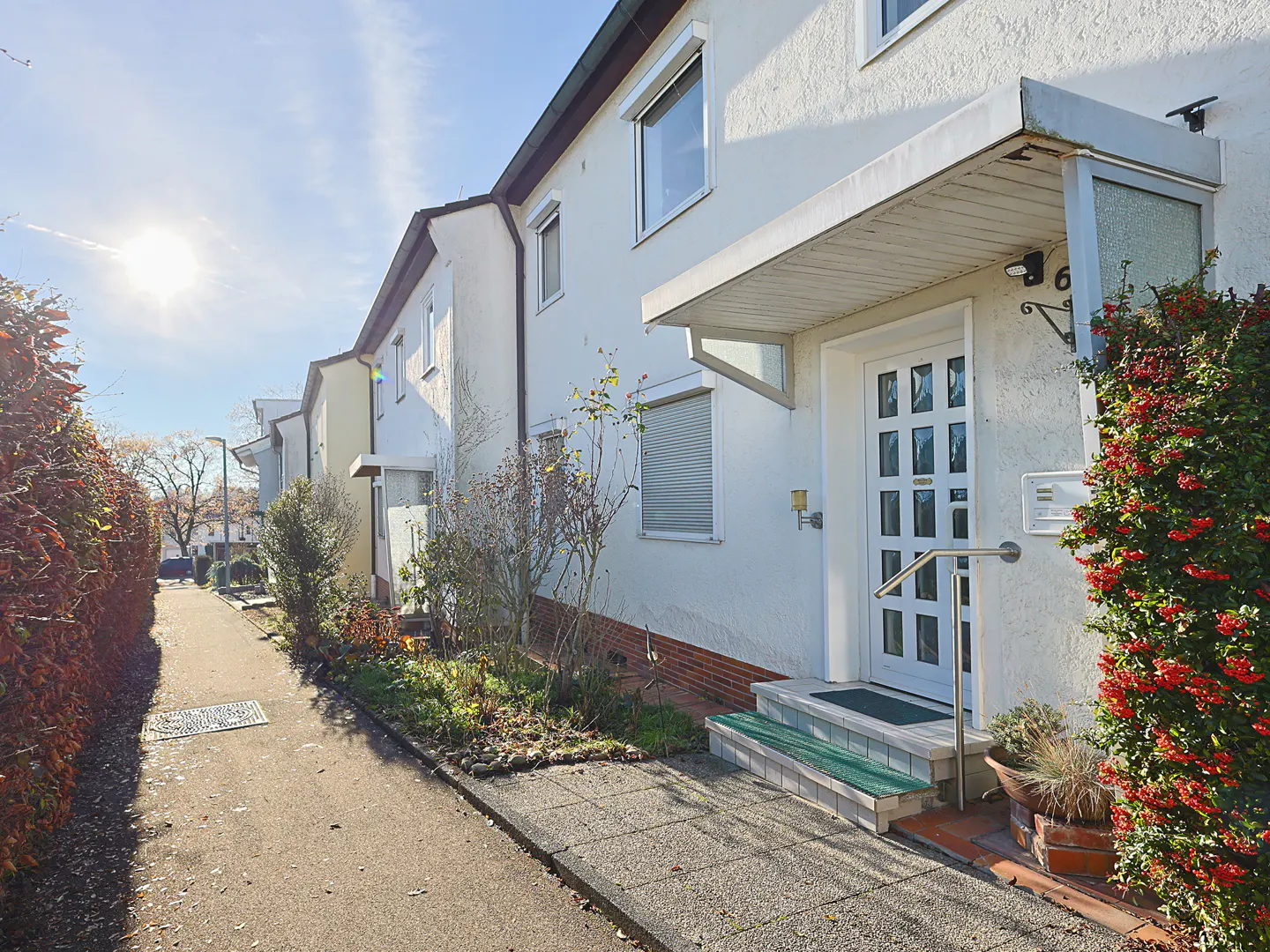 Exterior view of a row of white townhouses with a sidewalk, greenery, and a sunny sky. The front door has a small awning.