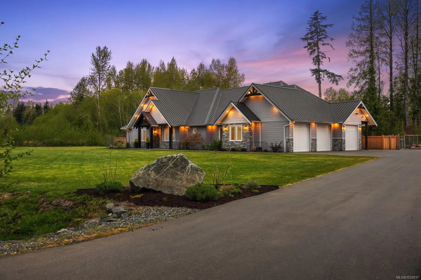 A gray house with a dark roof sits on a green lawn under a twilight sky. A large rock sits near the driveway.