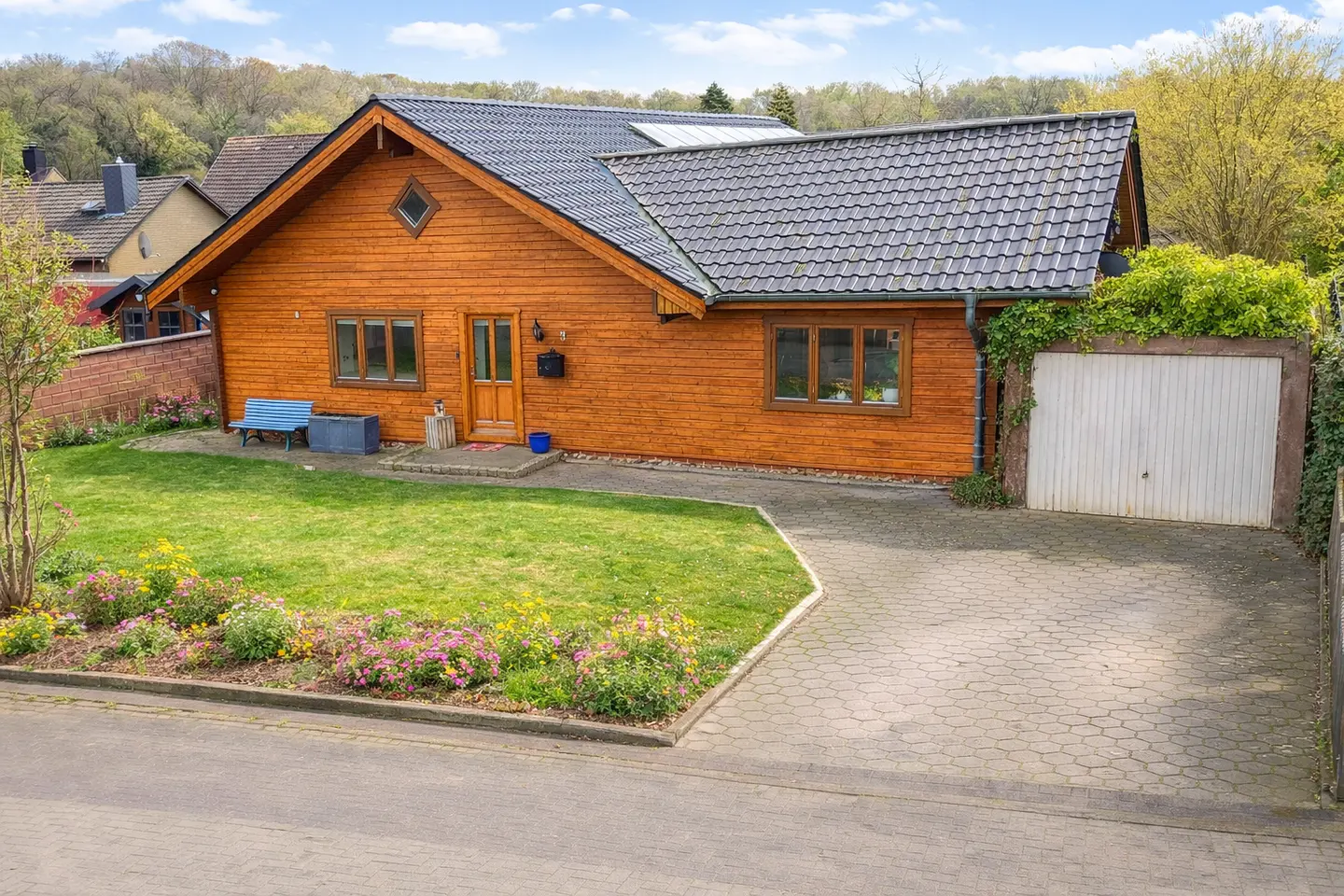 Exterior view of a one-story wood house with a gray roof, a green lawn, and a paved driveway.