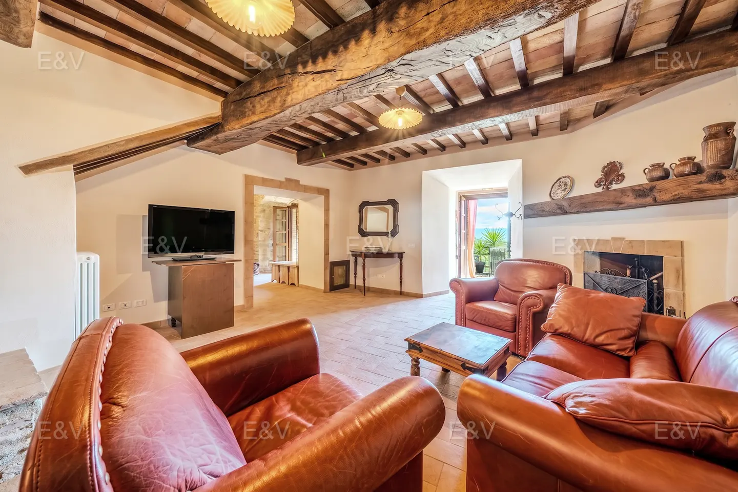 Rustic living room with terracotta floors, exposed wooden ceiling beams, leather sofas, and a fireplace.
