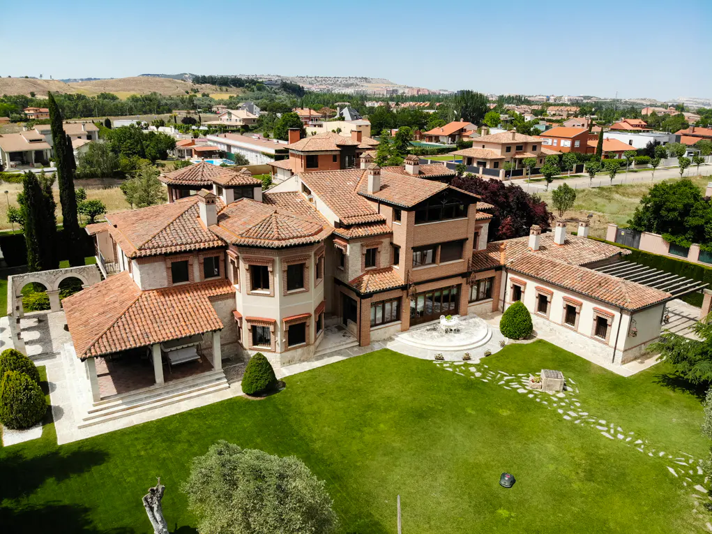 Aerial view of a large Mediterranean-style villa with a terracotta roof, expansive green lawn, and a suburban backdrop.