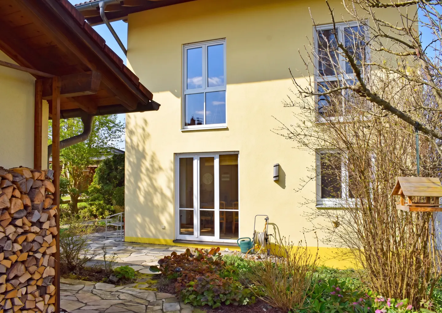 Exterior view of a yellow two-story house with white windows and a stone patio. A woodpile is visible on the left.