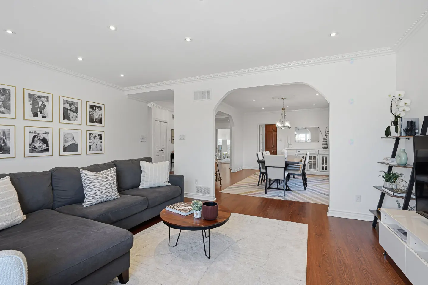 Bright living room with a gray sectional sofa, round wood coffee table, and gallery wall of framed photos. Dining room visible through an archway.