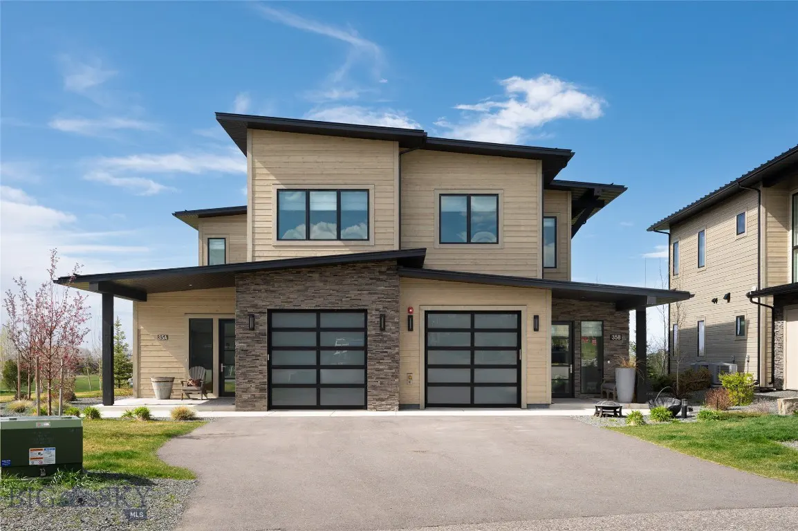 Two-story modern home with tan siding, stone accents, and black trim under a blue sky. Two-car garage with glass doors. Paved driveway.