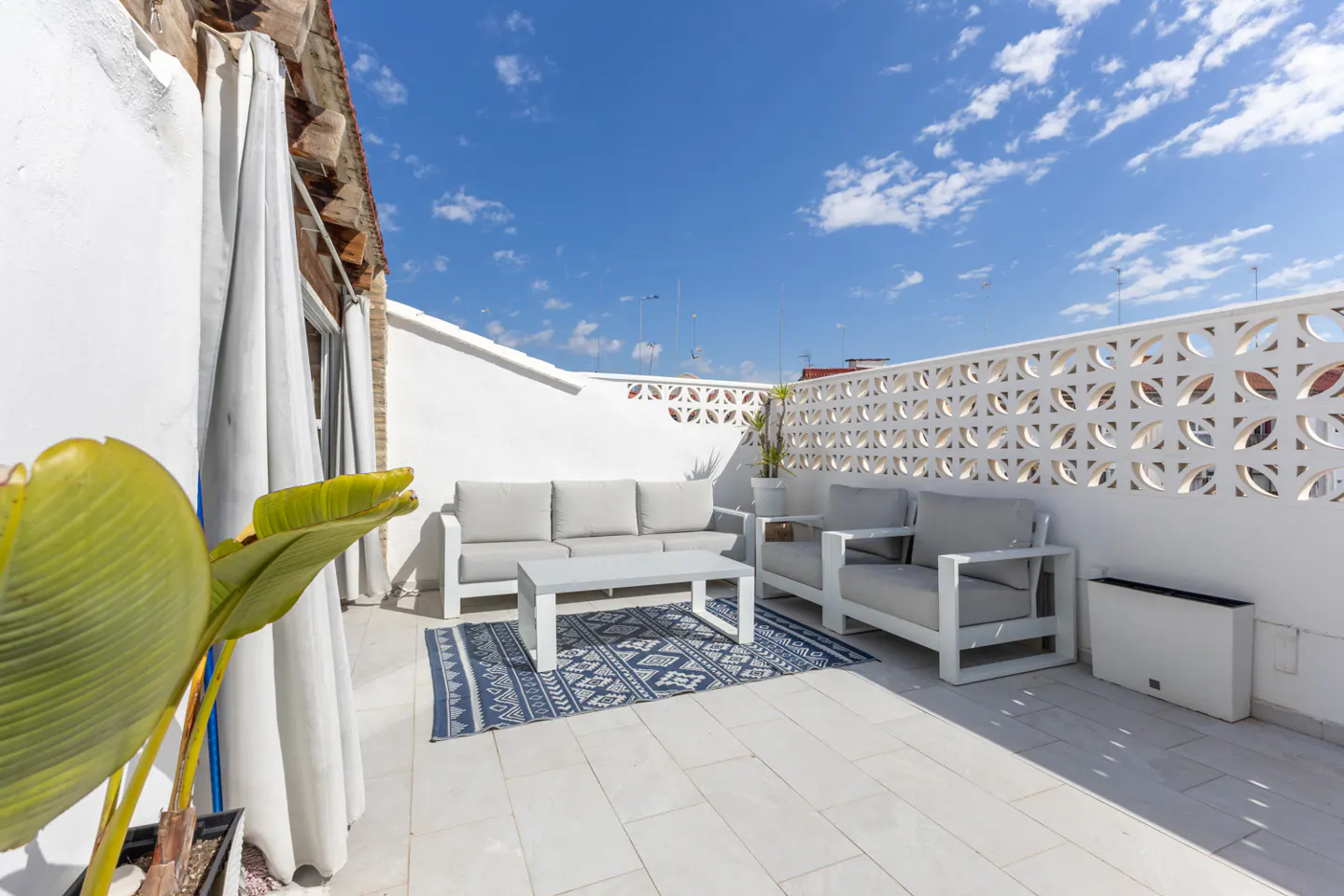 Bright rooftop patio with white walls, blue patterned rug, and gray outdoor furniture under a blue sky.