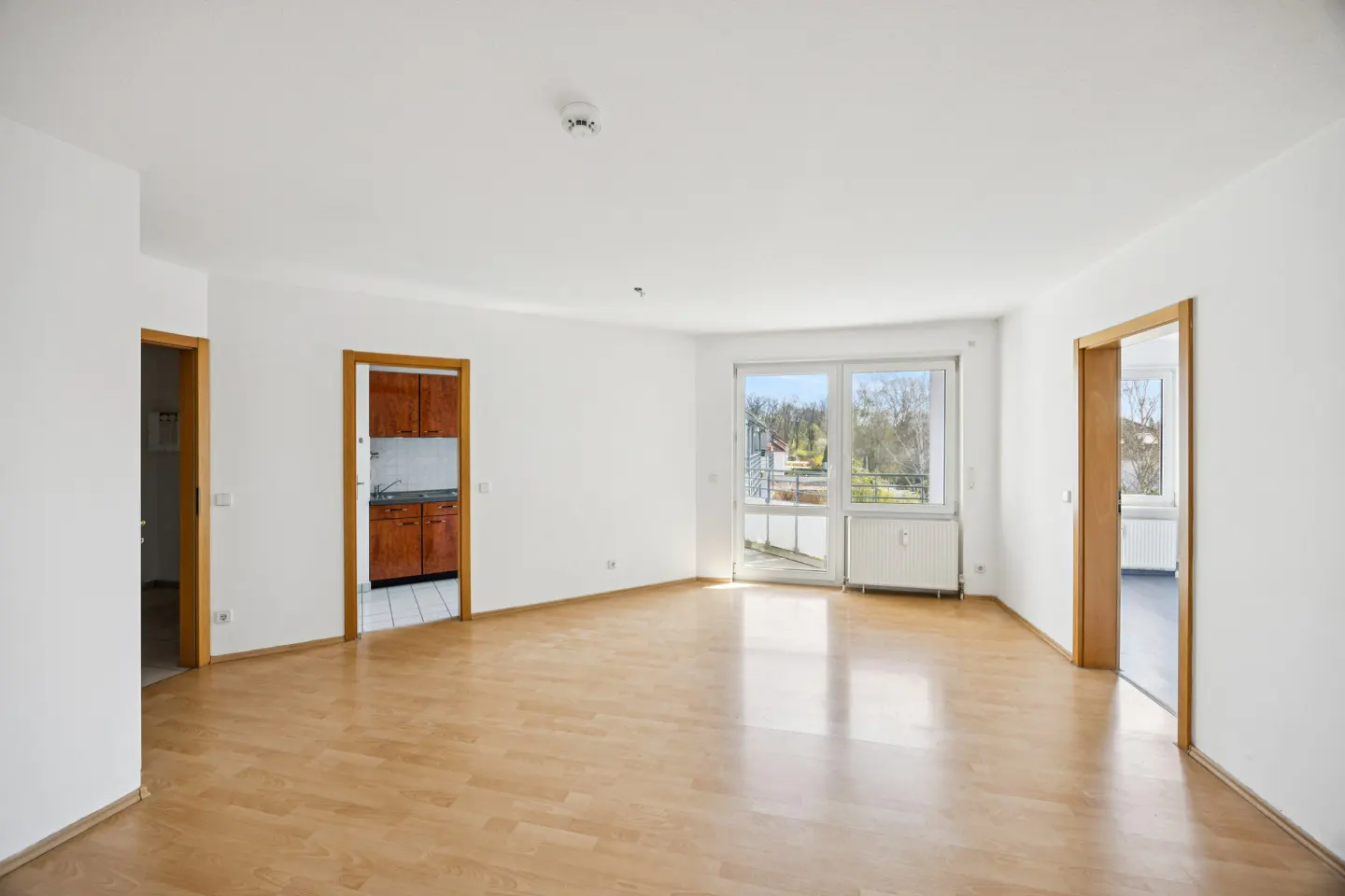 Bright, empty room with wood floors, white walls, and three doorways. One doorway leads to a kitchen with wood cabinets. Balcony with a view.