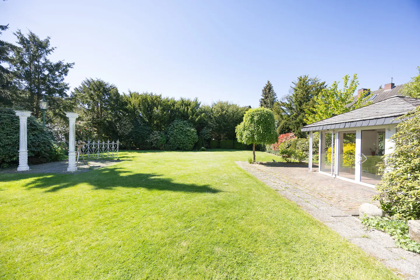 A lush green lawn with trees, white columns, and a sunroom with glass doors on a sunny day.