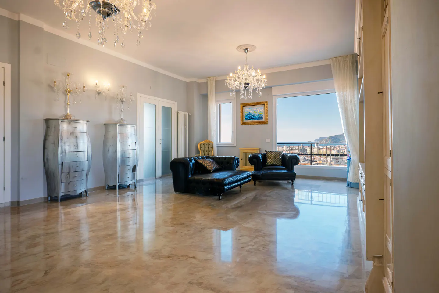 A bright living room with marble floors, black leather seating, and a large window overlooking a coastal city. Chandeliers hang from the ceiling.