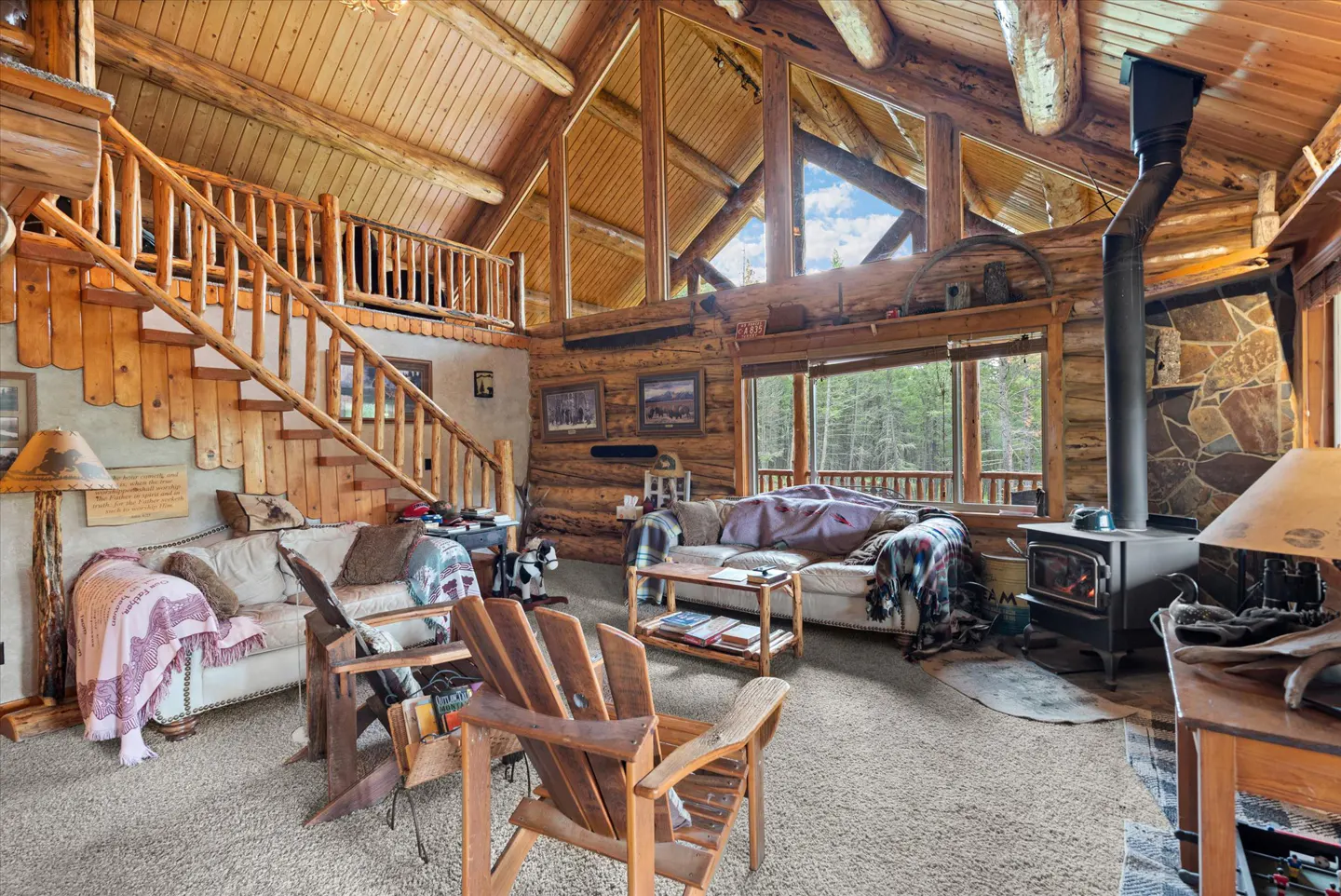 Rustic log cabin interior with wood walls, high ceilings, a stone fireplace, and large windows overlooking a forest.