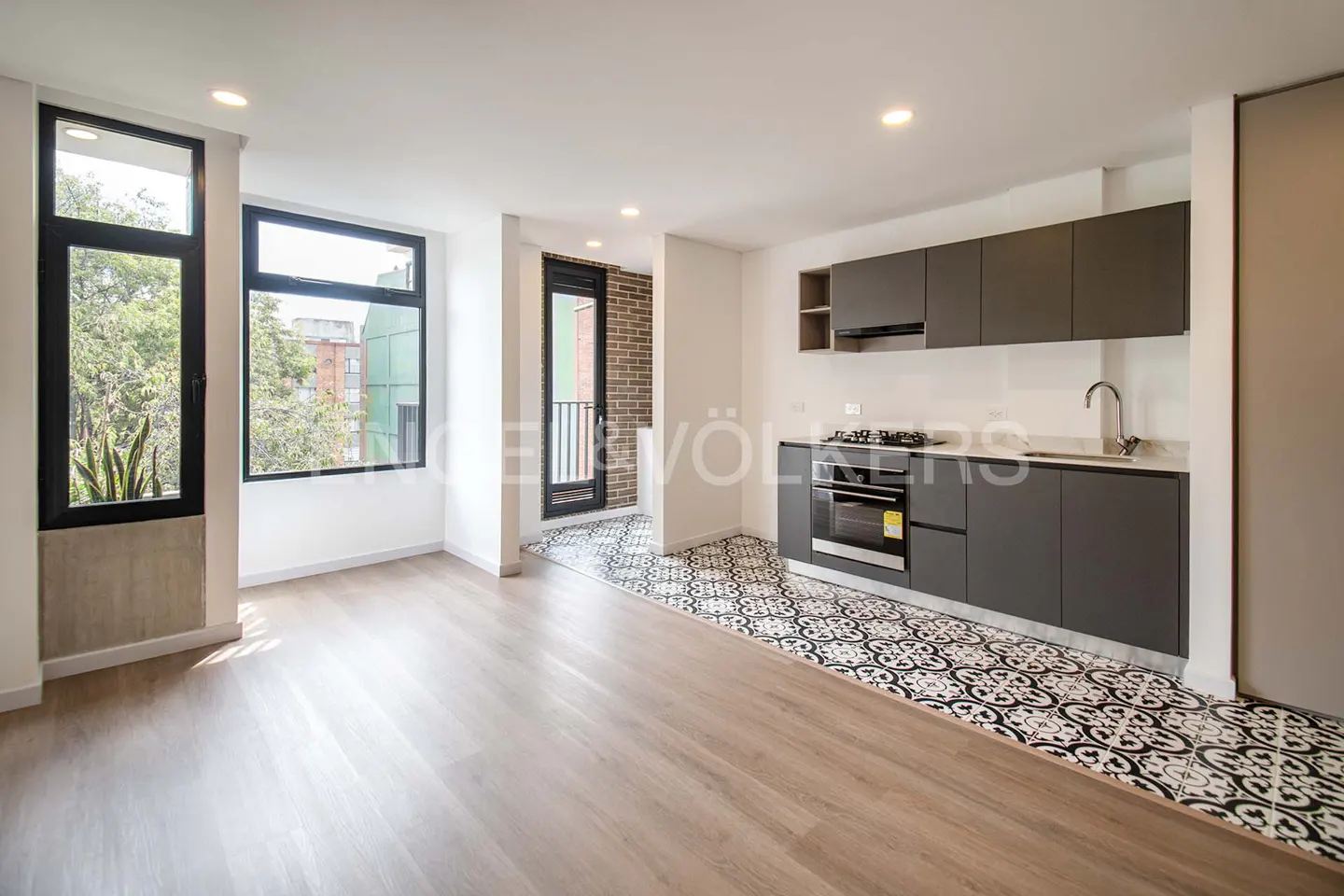 Bright, modern apartment with wood floors, black-framed windows, and a gray kitchen with patterned tile flooring.